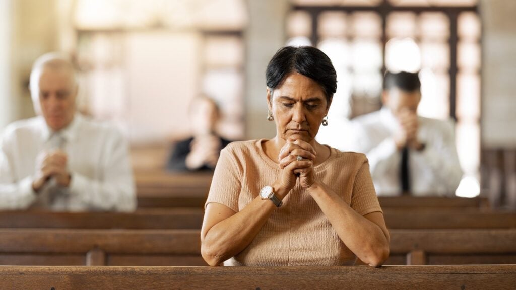A middle-aged woman praying in a church with others praying behind her.